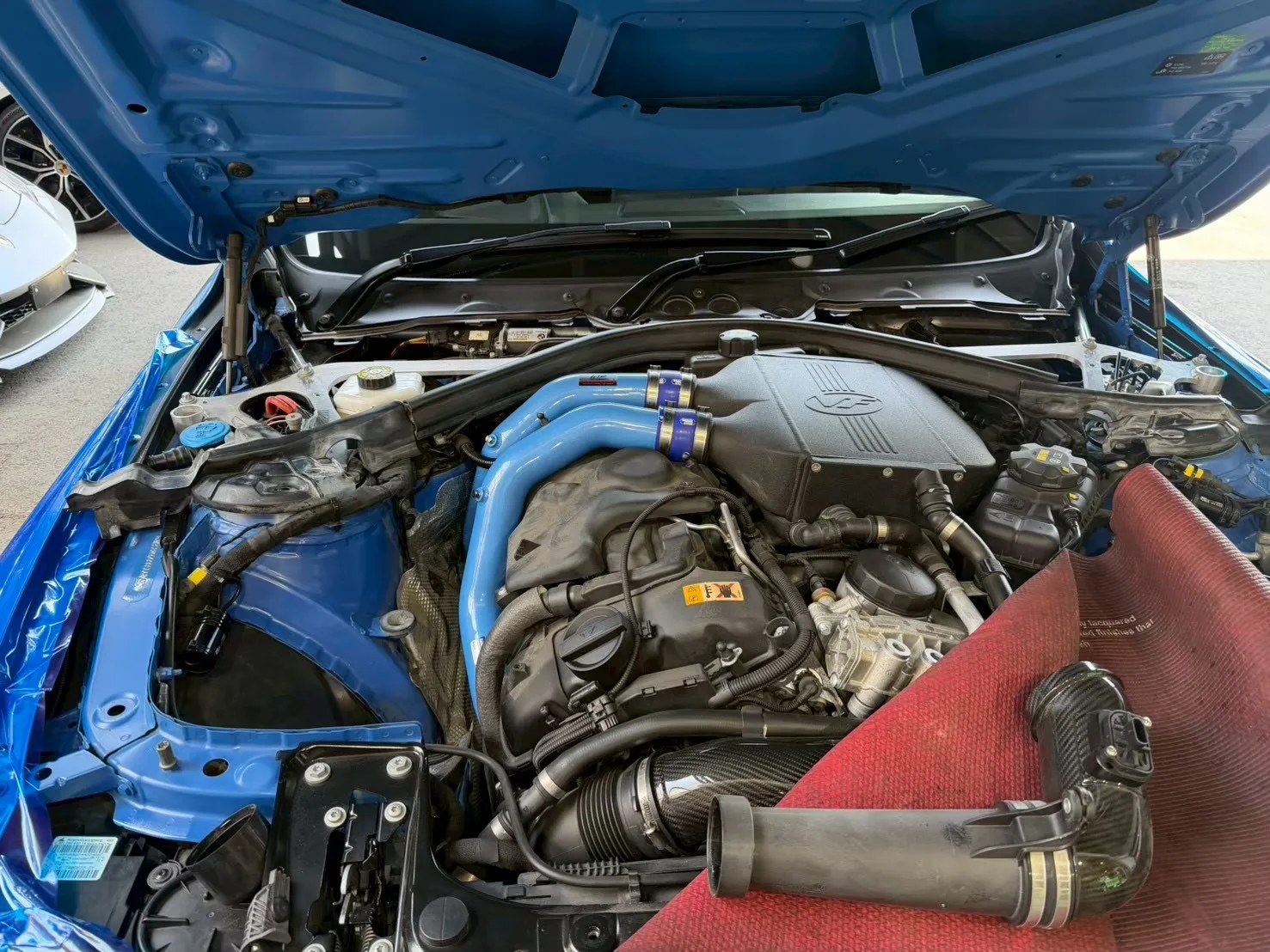 bmw-m3-charge-pipe-replacement-3 – mbFAST Tuning Open hood view of a blue car showing the engine bay with blue intake pipes and an oversized airbox at the center