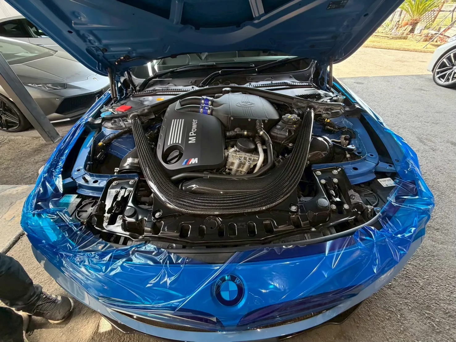 Blue BMW with hood open, showing a high-performance M Power engine and braided intake ducts in the foreground, battle-blue exterior wrap visible around the engine bay.