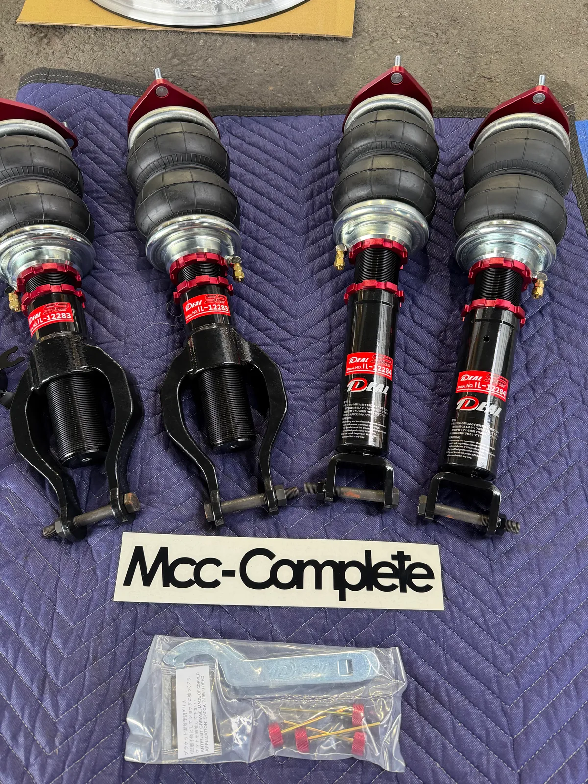gtr-exhaust-airsuspension-3 – mbFAST Tuning Set of four adjustable coilover shock absorbers with red mounting collars laid out on a purple quilted mat. MCC-Complete label visible.