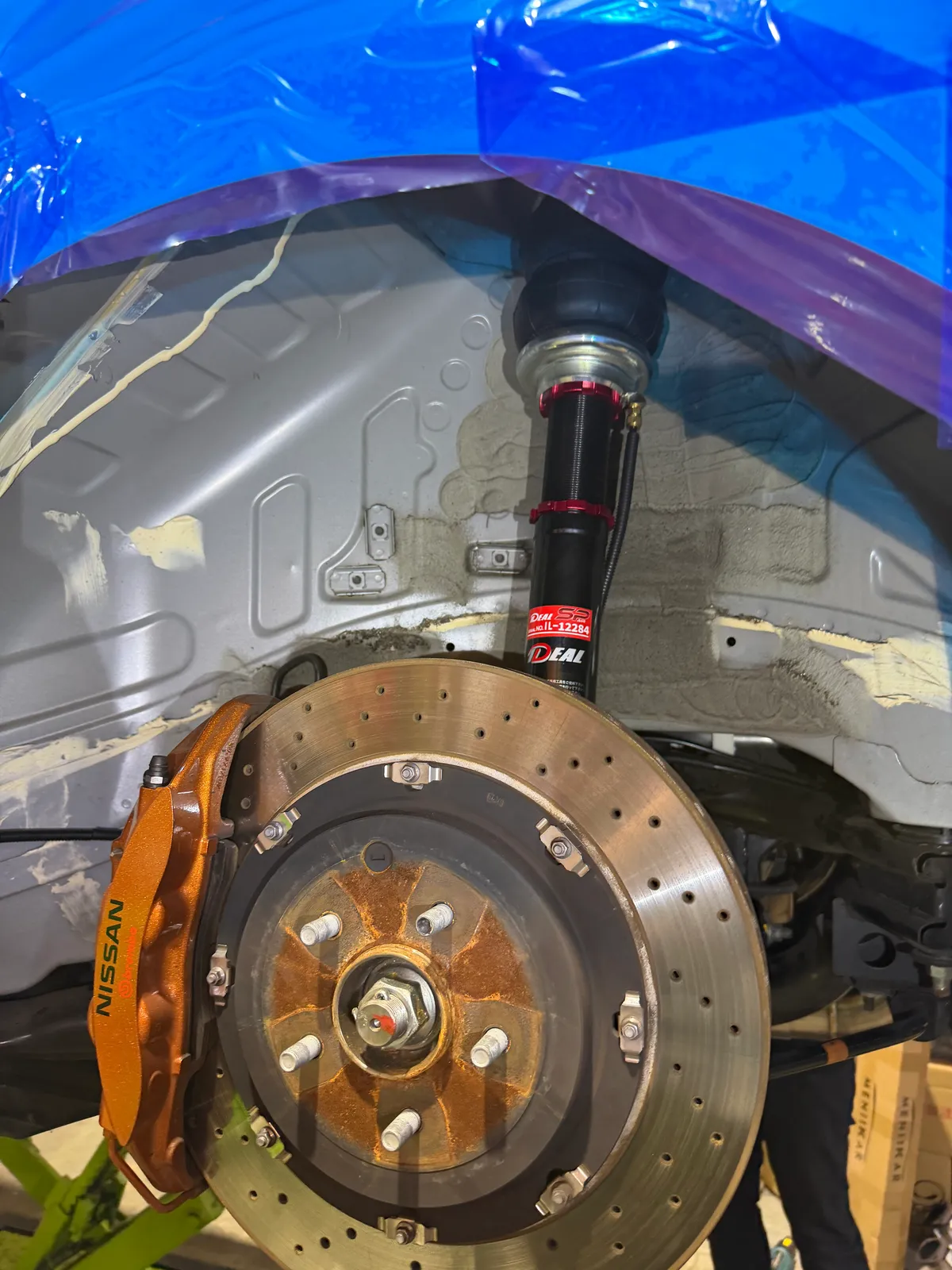 gtr-exhaust-airsuspension-6 – mbFAST Tuning Front brake assembly of a Nissan car: drilled rotor, orange caliper, and wheel hub visible with a black coilover suspension above in a workshop.