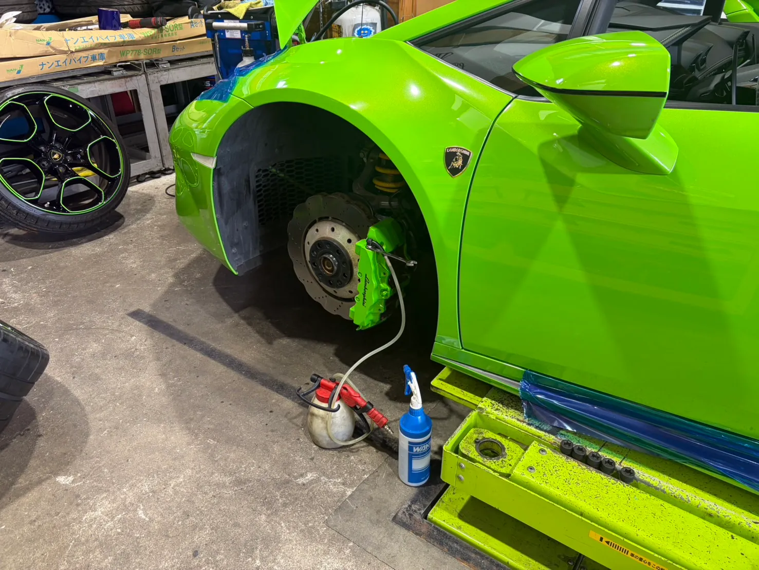huracan-caliper-coating-4 – mbFAST Tuning Lamborghini on a lift with the front wheel removed, exposing neon-green brake caliper and rotor in a workshop.