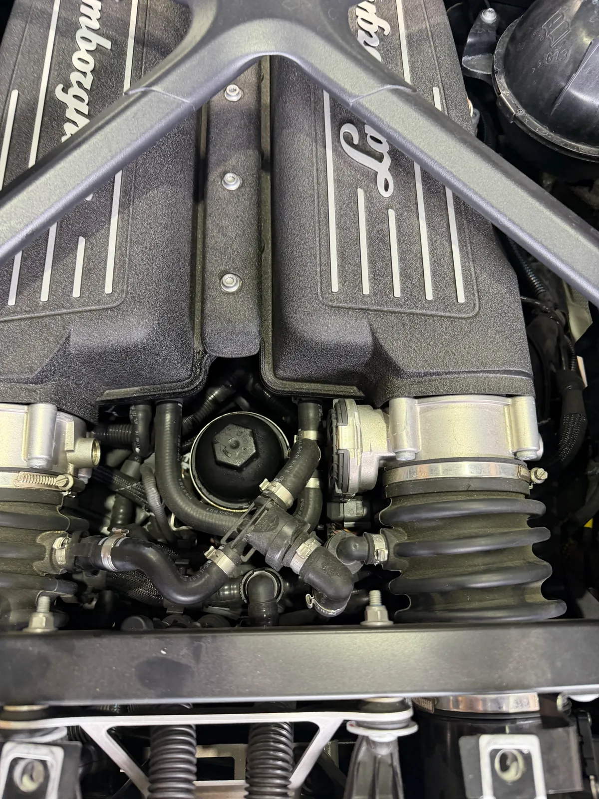 huracan-oil-change-10 – mbFAST Tuning Close-up of a Lamborghini V8 engine showing black valve covers with silver stripes and branding, with various hoses and intake components around them.