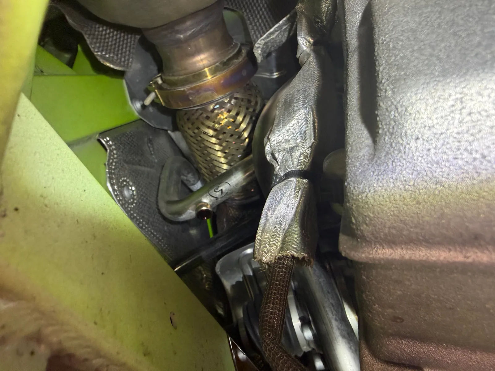huracan-oil-change-8 – mbFAST Tuning Close-up of a braided exhaust flex pipe and metal joint inside a vehicle’s undercarriage near a green chassis, showing heat shielding and wiring.